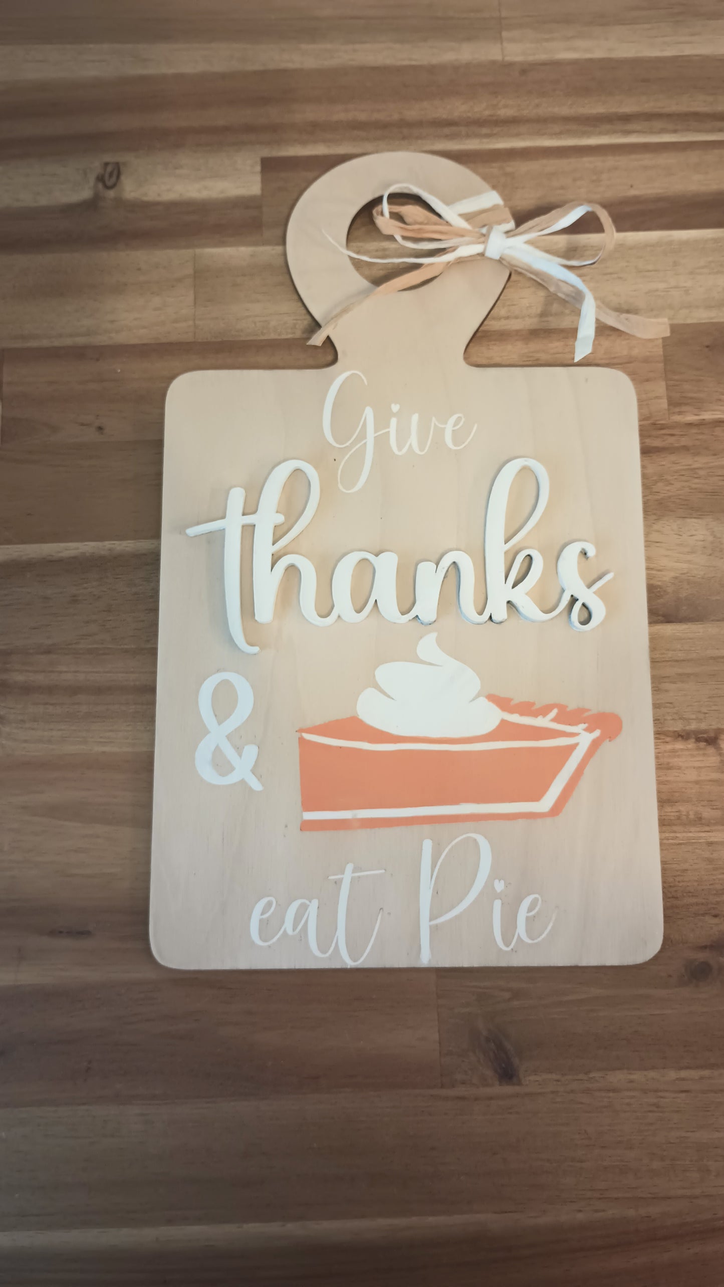 A decorative wooden sign shaped like a cutting board with a raffia bow at the top. The sign features the phrase "Give thanks & eat pie" in a mix of white and beige script and block letters. Below the text is an illustration of a slice of pumpkin pie topped with whipped cream. The sign has a warm, rustic, autumn-themed design.