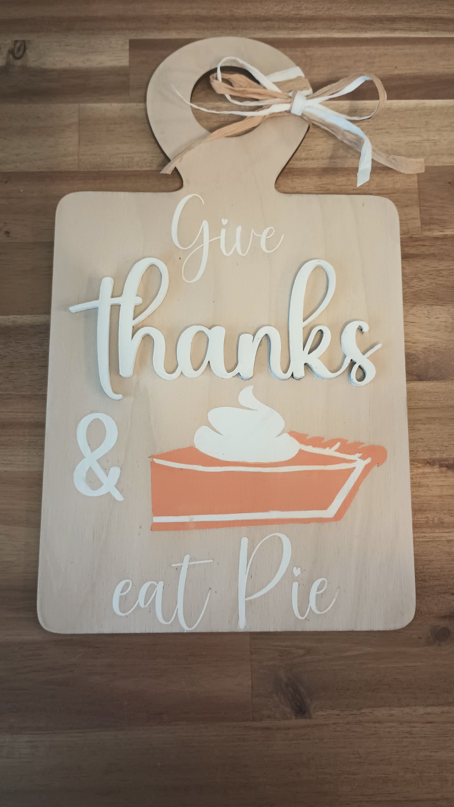 A decorative wooden sign shaped like a cutting board with a raffia bow at the top. The sign features the phrase "Give thanks & eat pie" in a mix of white and beige script and block letters. Below the text is an illustration of a slice of pumpkin pie topped with whipped cream. The sign has a warm, rustic, autumn-themed design.