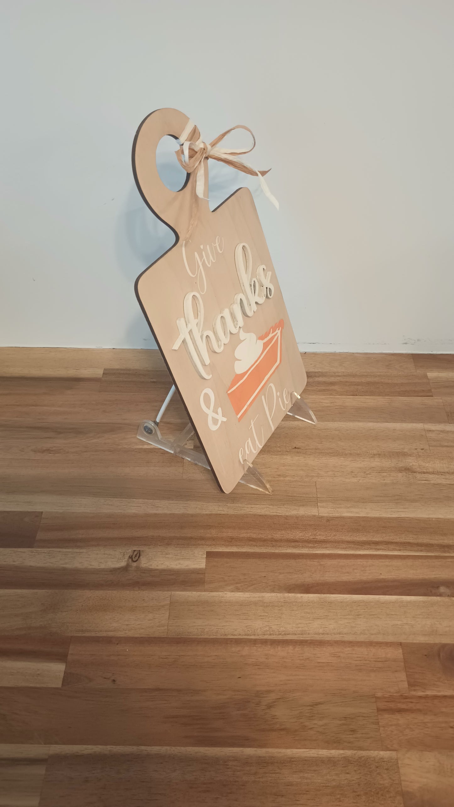 A decorative wooden sign shaped like a cutting board with a raffia bow at the top. The sign features the phrase "Give thanks & eat pie" in a mix of white and beige script and block letters. Below the text is an illustration of a slice of pumpkin pie topped with whipped cream. The sign has a warm, rustic, autumn-themed design.