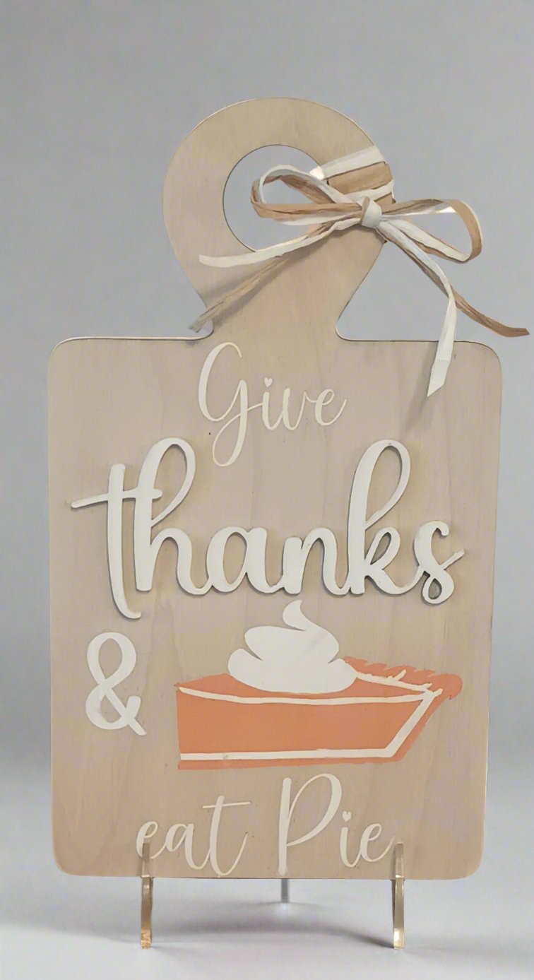 A decorative wooden sign shaped like a cutting board with a raffia bow at the top. The sign features the phrase "Give thanks & eat pie" in a mix of white and beige script and block letters. Below the text is an illustration of a slice of pumpkin pie topped with whipped cream. The sign has a warm, rustic, autumn-themed design.