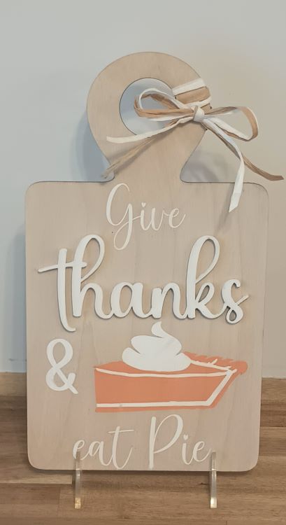 A decorative wooden sign shaped like a cutting board with a raffia bow at the top. The sign features the phrase "Give thanks & eat pie" in a mix of white and beige script and block letters. Below the text is an illustration of a slice of pumpkin pie topped with whipped cream. The sign has a warm, rustic, autumn-themed design.