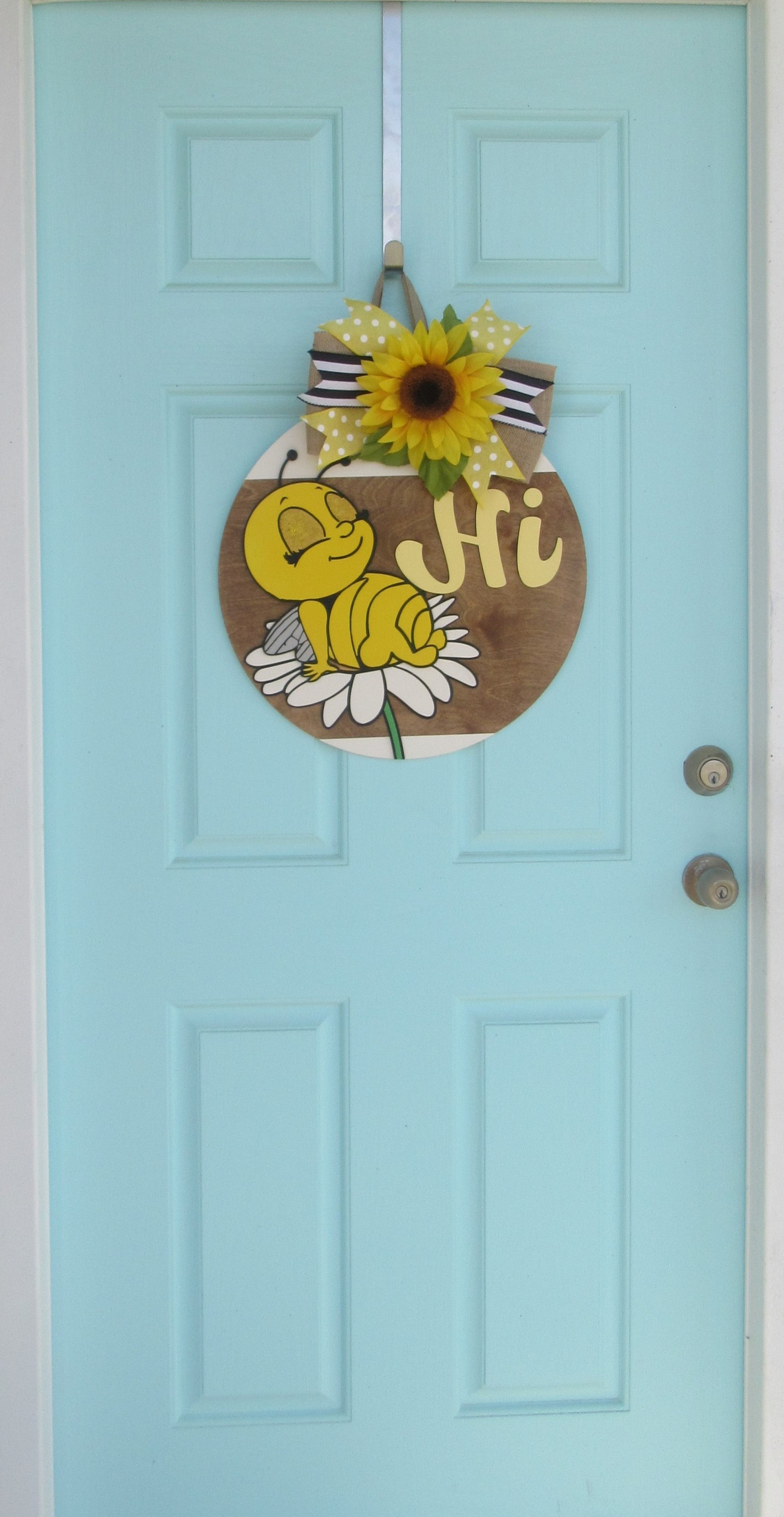 Bee-themed wooden door hanger with “Hi” greeting and sunflower bow displayed on a light blue front door