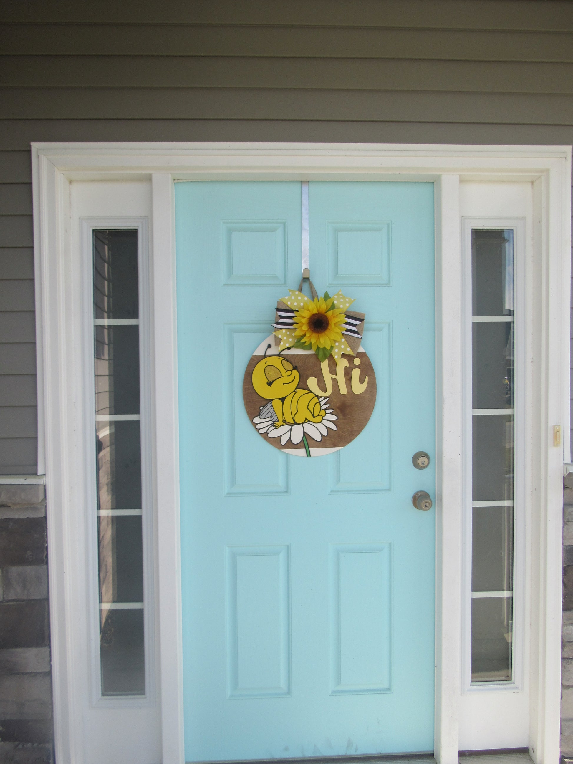 Bee-themed wooden door hanger with “Hi” greeting and sunflower bow displayed on a light blue front door