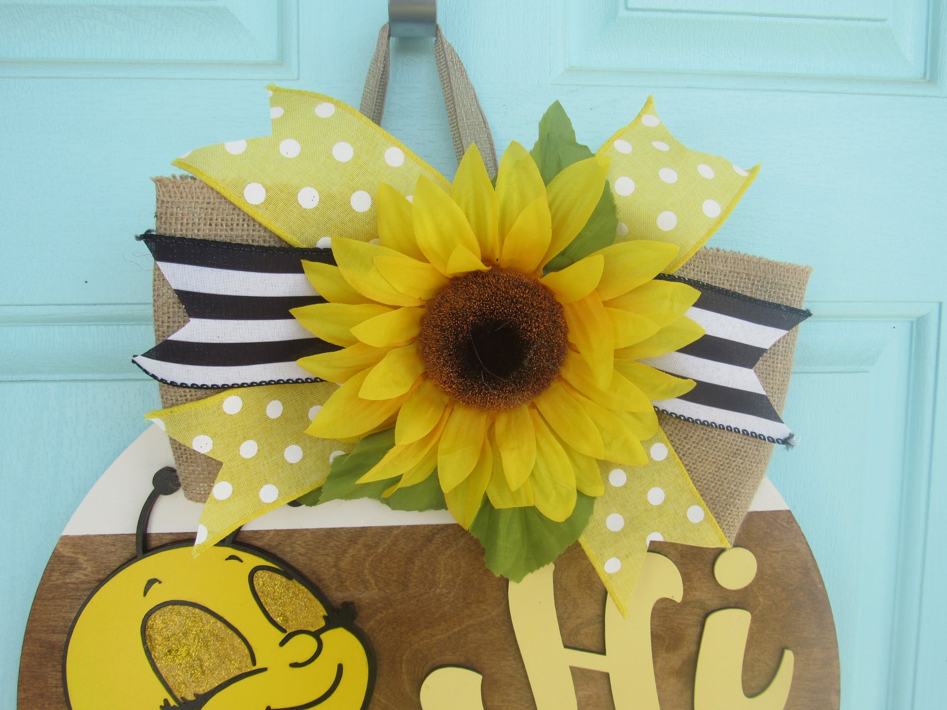 Bee-themed wooden door hanger with “Hi” greeting and sunflower bow displayed on a light blue front door