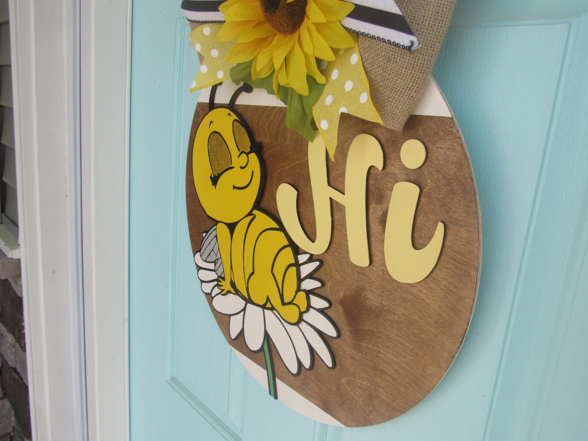 Bee-themed wooden door hanger with “Hi” greeting and sunflower bow displayed on a light blue front door
