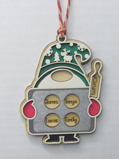 A close-up of a wooden Christmas ornament shaped like a gnome. The gnome wears a green and white hat decorated with forest silhouettes, including a deer and trees. It has red mittens and holds a rectangular silver tray with four circular cutouts, each engraved with a name: "James," "Tanya," "Lucas," and "Emily." A rolling pin on the right side of the gnome is engraved with the year "2023." The ornament is placed on a plain white background.