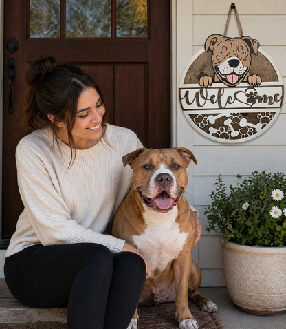 Woman sitting with a pit bull dog in front of a 'Welcome' sign with a dog design on a wooden door.