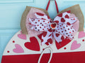 Close-up of a wooden heart door hanger, showcasing red and pink hand-painted hearts, a layered polka dot bow, and sparkly red heart charm.