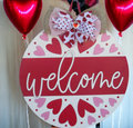 Front view of a wooden heart Valentine door sign with pink and red heart patterns, a 'Welcome' greeting, and festive bow detail, perfect for gifting.