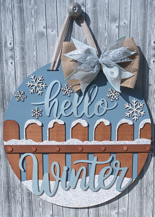 Handcrafted Hello Winter door hanger featuring snowflakes, glitter accents, a burlap bow, and a snowy picket fence, perfect for winter front door decor.