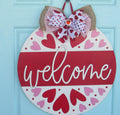 Wooden Valentine’s Day door sign with a vibrant red 'Welcome' text, 18 inch diameter, handcrafted with heart patterns and a festive bow accent